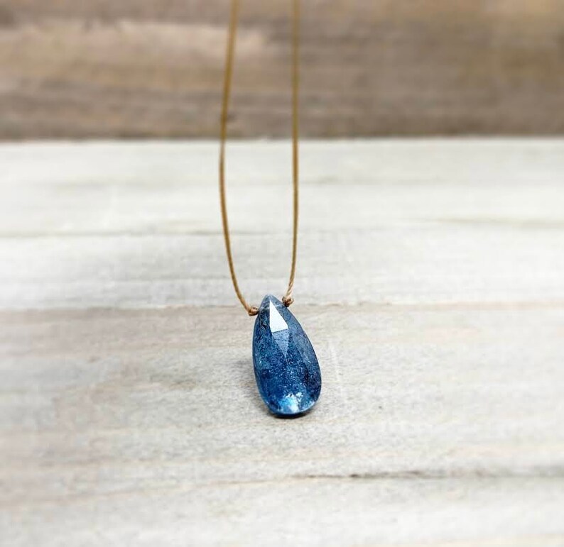 May include: A teardrop-shaped blue gemstone pendant on a thin brown cord necklace.