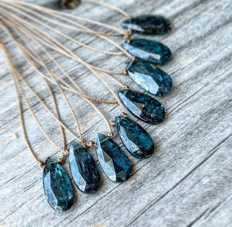 May include: A set of nine teal blue gemstone pendants on brown cords. The pendants are faceted and have a teardrop shape.