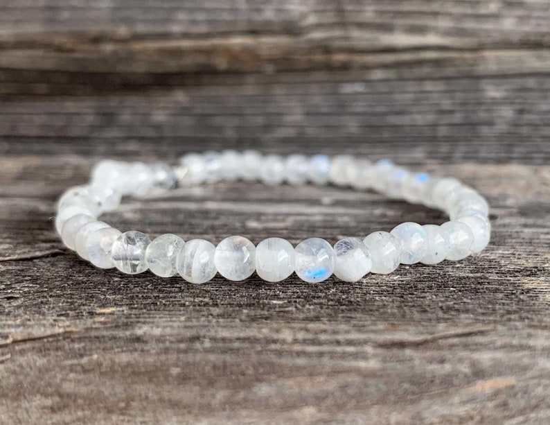 May include: A white moonstone bracelet with a silver clasp. The bracelet is made of round beads and is lying on a wooden surface.