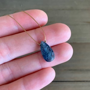 May include: A teardrop-shaped blue gemstone pendant on a brown cord necklace.