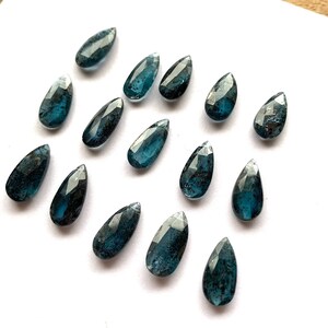May include: A collection of 15 teardrop-shaped blue apatite gemstone beads. The beads are faceted and have a dark blue color with some black inclusions.