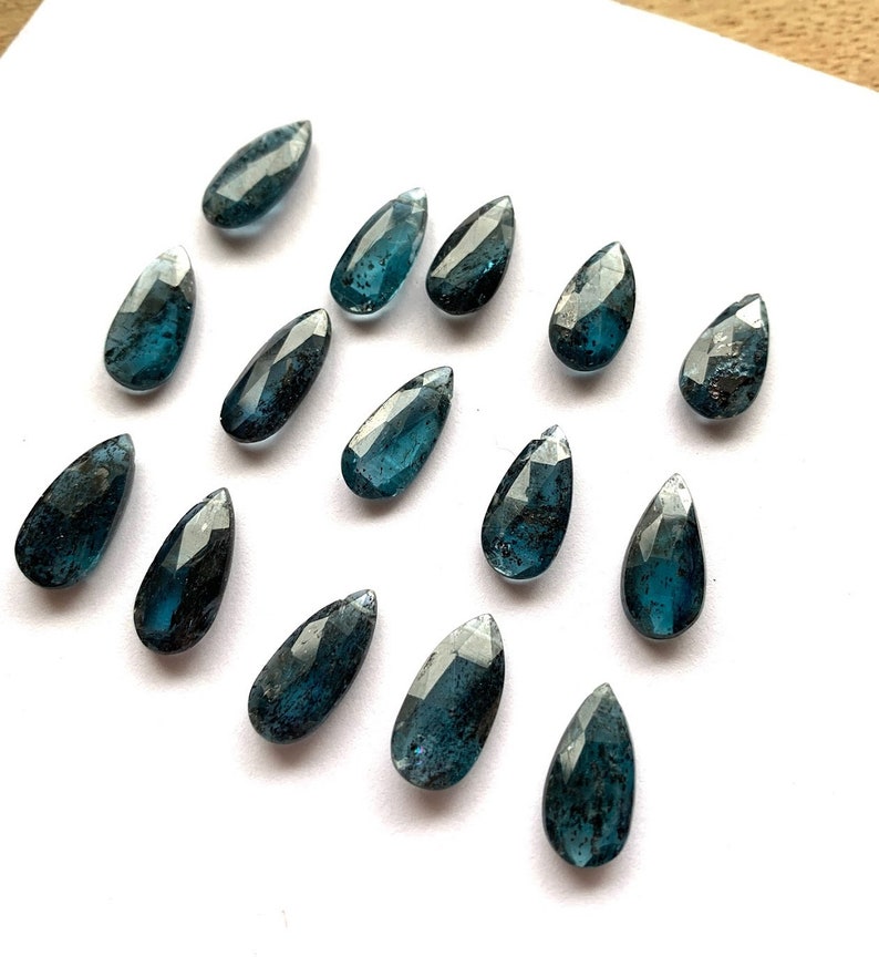 May include: A collection of 15 teardrop-shaped blue apatite gemstone beads. The beads are faceted and have a dark blue color with some black inclusions.
