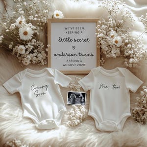Puede incluir: Anuncio de embarazo con dos bodies blancos para bebés, uno con la inscripción "Coming Soon" y el otro "Me too!". Un letrero enmarcado dice "We've been keeping a little secret, anderson twins, arriving August 202X". La imagen incluye una ecografía y flores.