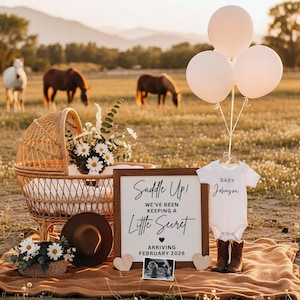 Könnte beinhalten: Eine rustikale Szene im Freien mit einer Babyankündigung. Ein Holzschild liest: "Saddle Up! We've been keeping a little secret, arriving February 2026." Ein Baby-Body, Cowboy-Stiefel und Ballons sind ausgestellt. Eine Weidenkorb-Wiege und Pferde im Hintergrund.