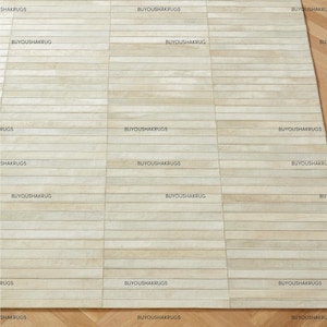 Op de afbeelding: Een rechthoekig tapijt met een lichtbeige, horizontaal gestreept patroon. Het tapijt heeft een textuur en is gemaakt van een zacht materiaal. De woorden "BUYOUSHAKRUGS" en "SHAKRUGS" staan op het tapijt.