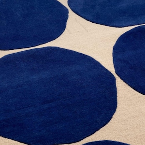 May include: A rug featuring a repeating pattern of large, solid dark blue circular shapes set against a neutral, light beige background. The surface of the rug has a textured, woven appearance with distinct color contrast between the blue circles.