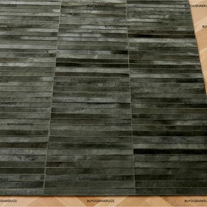 May include: A rectangular rug with a patchwork design. The rug is composed of horizontal strips in shades of gray and black. The texture appears to be soft and plush. The words "BUYOUSHAKRUGS" are printed on the rug.