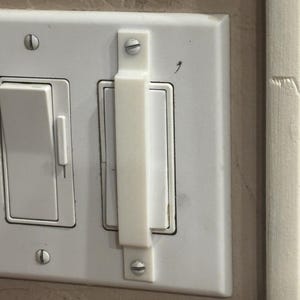 May include: A white light switch cover with a white plastic light switch extender attached. The cover has two switches and is mounted on a beige wall. The extender is secured with two screws.