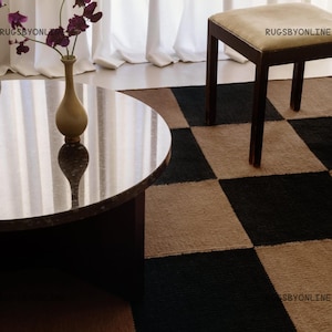 Könnte beinhalten: Eine moderne Wohnzimmerszene mit einem runden Couchtisch, einem karierten Teppich und einem kleinen Hocker. Der Teppich hat ein schwarz-braunes Schachbrettmuster. Eine Vase mit lila Blumen steht auf dem Tisch. Der Hocker hat ein beiges Kissen und dunkelbraune Beine.