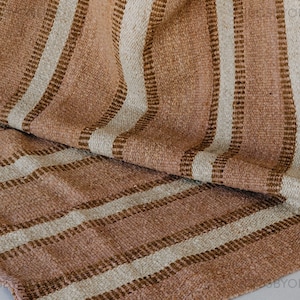 Puede incluir: Textil tejido con un patrón de rayas en tonos marrón y crema. La tela está doblada, revelando la textura y el diseño. Las rayas verticales están espaciadas uniformemente, creando un aspecto simple pero elegante. El textil parece estar hecho de fibras naturales.