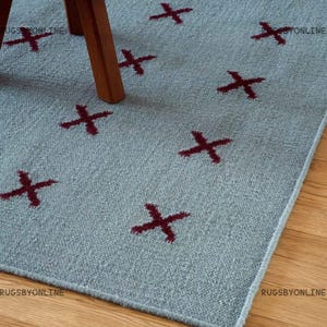 Op de afbeelding: Een lichtblauw tapijt met een herhalend patroon van bordeauxrode kruisen. Het tapijt is gemaakt van een textuurrijk materiaal en ligt op een houten vloer. Een houten stoelpot is zichtbaar in de afbeelding.