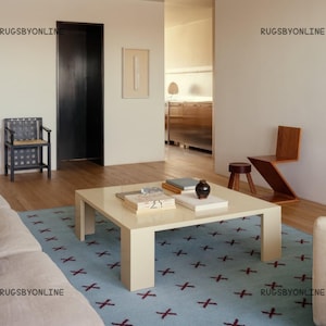 Può includere: Un soggiorno moderno con un tappeto azzurro con un motivo a croce bordeaux ripetuto. Un tavolino quadrato color crema contiene libri e oggetti decorativi. La stanza comprende un divano, sedie e pavimenti in legno. Le pareti sono dipinte in un colore neutro.