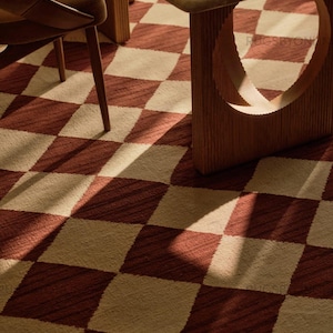 May include: A floor rug featuring a repeating diamond pattern in alternating shades of deep terracotta red and cream. The textile surface displays a visible woven texture, with the text RUGSBYON visible in the upper right corner of the rug.