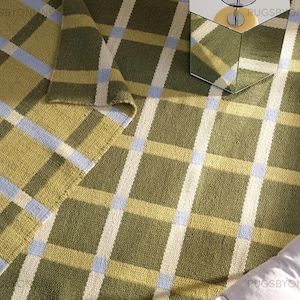 May include: A green and yellow plaid rug with blue accents. The rug is made of a woven material and has a geometric pattern. A hexagonal mirrored side table with a lamp is on the rug.