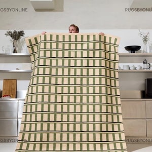 May include: A large rectangular rug with a repeating pattern of green and black stripes on a beige background. The rug is being held up by a person, showcasing its size and design. The rug is suitable for a living room or bedroom.
