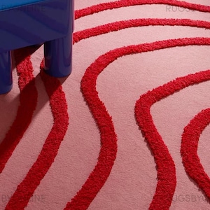 Peut inclure: Tapis rose avec des rayures rouges ondulées en relief. Une petite table bleue avec un plat décoratif blanc est visible en haut à gauche. Le tapis a un aspect texturé et une bordure à franges.
