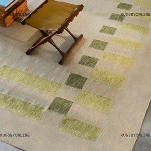 May include: Beige area rug with a geometric pattern in shades of green and yellow. A folding wooden stool with a tan leather seat is in the foreground. The rug has the text "RUGSBYONLINE" in the corners.