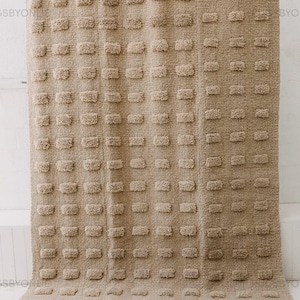 May include: A beige rectangular rug with a textured, tufted pattern. The rug features a repeating design of raised, rectangular shapes across its surface. The neutral color and simple design make it suitable for various interior styles.