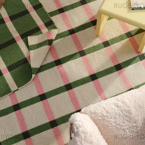 May include: A woven rug with a plaid pattern in green, pink, black, and cream. The rug is partially folded, revealing the underside. A small yellow table and a fluffy white pet bed are also visible.