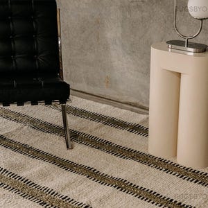 Puede incluir: Una alfombra a rayas de color crema con rayas negras y verde oliva. Una silla de cuero negro con estructura cromada está a la izquierda. Una mesa auxiliar beige con una lámpara cromada está a la derecha.