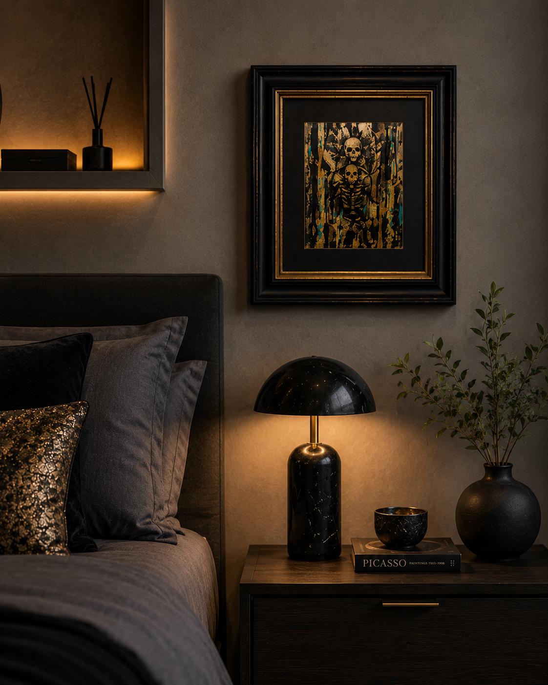 May include: A black marble table lamp with a dome-shaped shade and a gold-toned neck, positioned on a dark wooden nightstand next to a black vase with green foliage, a small black bowl, and a book titled Picasso, beneath a framed skull artwork.