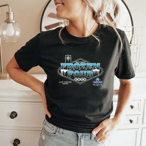 Op de afbeelding: Zwart T-shirt met de tekst "NCAA Men's Frozen Four 2020 Las Vegas Nevada" in blauw, wit en goud. Het ontwerp staat op de voorkant. Het shirt heeft een ronde hals en korte mouwen.