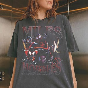 以下が含まれることがあります： チャコールグレーの半袖Tシャツで、様々なスーパーヒーローのポーズをとるマイルズ・モラレスのグラフィックプリントが施されています。上部には大きな白い文字で「MILES」、下部には大きな赤い文字で「MORALES」とプリントされています。