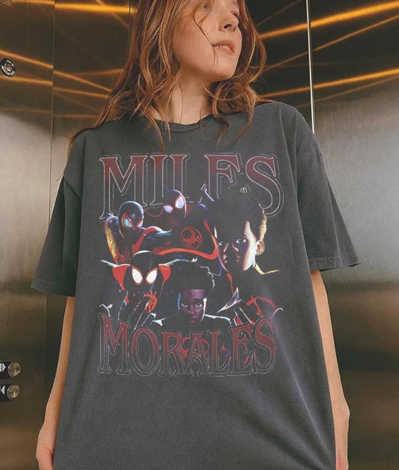 以下が含まれることがあります： チャコールグレーの半袖Tシャツで、様々なスーパーヒーローのポーズをとるマイルズ・モラレスのグラフィックプリントが施されています。上部には大きな白い文字で「MILES」、下部には大きな赤い文字で「MORALES」とプリントされています。