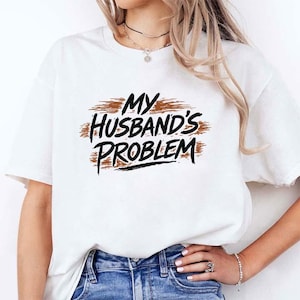 May include: White t-shirt with the text "My Husband's Problem" in black and brown brushstroke lettering. The shirt is a casual style, suitable for everyday wear.