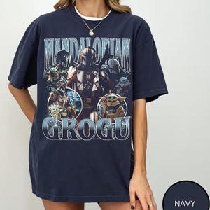 May include: A navy blue short-sleeve t-shirt featuring a graphic print of The Mandalorian and Grogu. The design includes the text "MANDALORIAN" at the top and "GROGU" at the bottom, with multiple character illustrations in a vintage-inspired style.