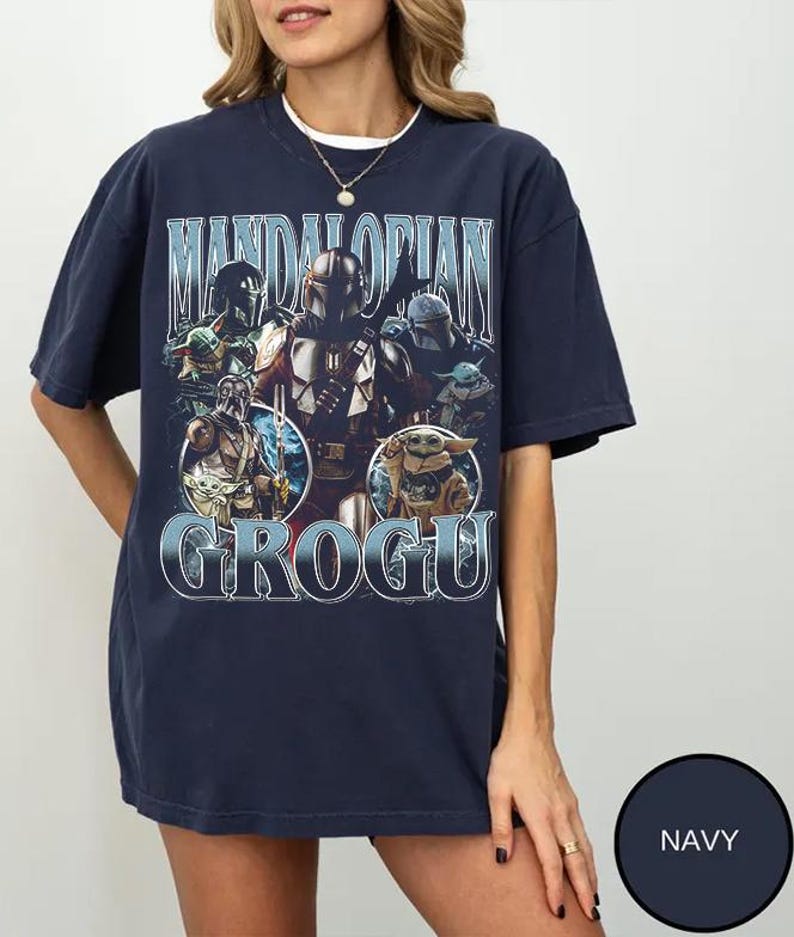 May include: A navy blue short-sleeve t-shirt featuring a graphic print of The Mandalorian and Grogu. The design includes the text "MANDALORIAN" at the top and "GROGU" at the bottom, with multiple character illustrations in a vintage-inspired style.