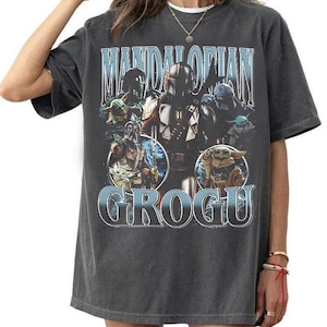 May include: A charcoal gray short-sleeve t-shirt featuring a graphic print of The Mandalorian and Grogu. The design includes the text "MANDALORIAN" at the top and "GROGU" at the bottom in light blue lettering, with multiple character illustrations.