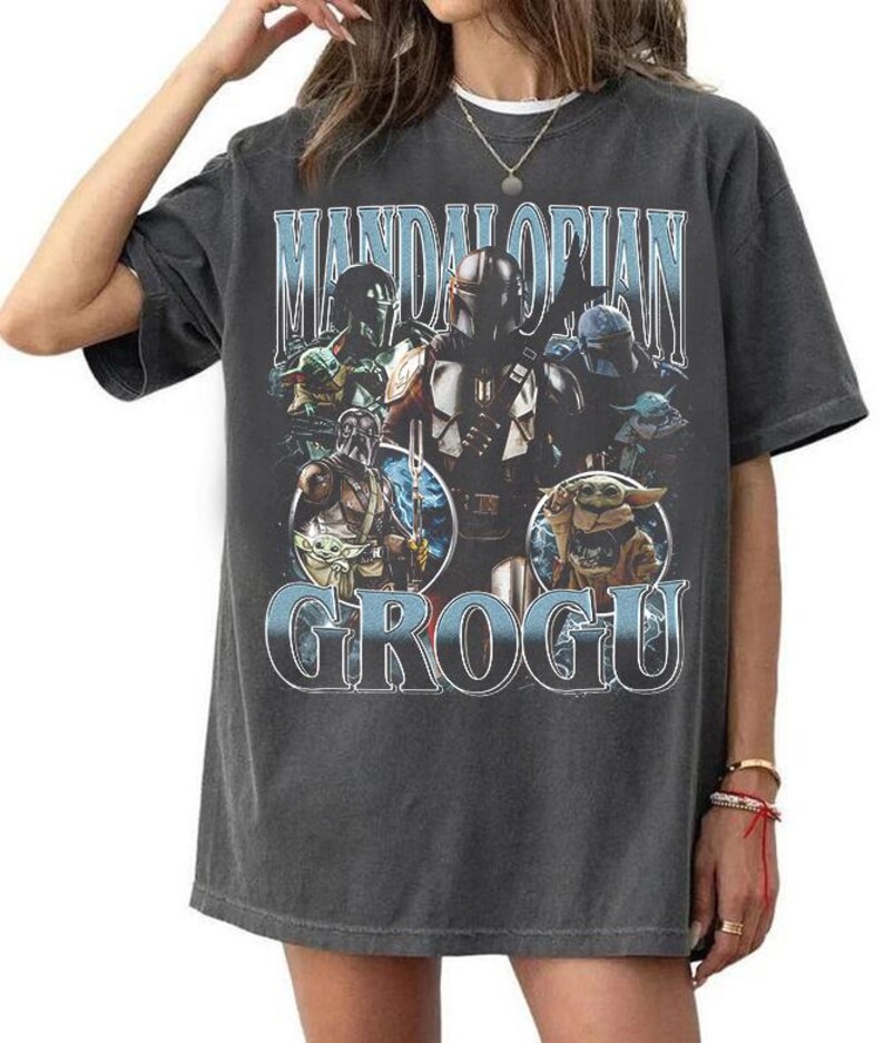 May include: A charcoal gray short-sleeve t-shirt featuring a graphic print of The Mandalorian and Grogu. The design includes the text "MANDALORIAN" at the top and "GROGU" at the bottom in light blue lettering, with multiple character illustrations.