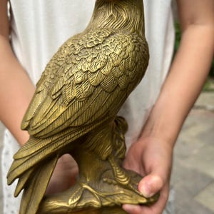 Può includere: Una statuetta di aquila in metallo color oro posata su un ramo testurizzato. La scultura presenta dettagliati motivi di piume incisi sulle ali e sul corpo, con uno sguardo acuto e un becco ricurvo, raffigurando l'uccello in posizione eretta.