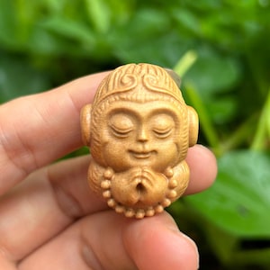 May include: A small, intricately carved wooden monkey figurine. The monkey is depicted with closed eyes, hands clasped in prayer, and a serene expression. The carving showcases detailed features and a smooth, polished finish. The figurine is held against a blurred green background.