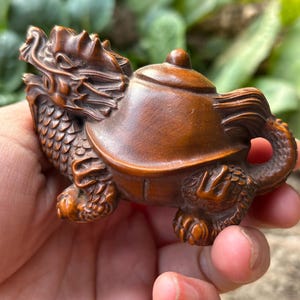 Op de afbeelding: Gedetailleerde houten sculptuur van een draak-schildpad hybride. De sculptuur is een theepot met een drakenkop, een schildpadschild en draakachtige voeten. Het hout heeft een warme bruine kleur en de gravure toont ingewikkelde details.