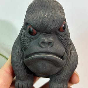 May include: A black gorilla figurine with red eyes. The gorilla has a stern expression and is sculpted with textured details. The figurine is held in a person's hand, showcasing its size and design. The gorilla's fur is textured, and the eyes are a striking red.