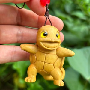 Op de afbeelding: Een lichtbruin houten beeldje van een schildpadachtig wezen met uitgestrekte armen en een glimlach. Het heeft gele ogen en is bevestigd aan een bruin koord met een rode kraal. Een hanger.