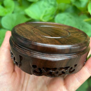 Puede incluir: Un soporte de exhibición de madera marrón oscuro con una parte superior circular y detalles decorativos tallados alrededor de la base. El soporte se sostiene en una mano, con follaje verde en el fondo.