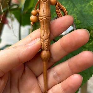 May include: A handcrafted wooden keychain featuring a carved, elongated design with intricate details. The keychain is a warm brown color, with a small loop at the end and a decorative cord with wooden beads. The background is blurred green foliage.