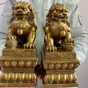 May include: A pair of golden lion statues, each on a decorative base. The lions have detailed manes and expressive faces, holding a ball and a cub. The bases feature intricate floral and geometric patterns, showcasing a traditional design.