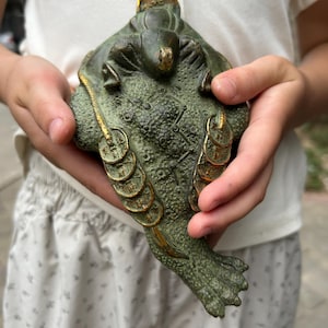May include: A dark green, textured metal sculpture of a three-legged toad with a bumpy back. The toad features gold-colored accents on its head and along the strings of circular coins draped over its sides and back, with a single coin on its head.