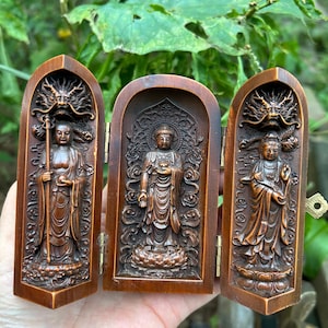 May include: A wooden triptych featuring carved figures of Buddha. The hinged panels open to reveal a central Buddha figure, flanked by two panels with additional figures and dragon motifs at the top. The wood has a warm, brown tone.