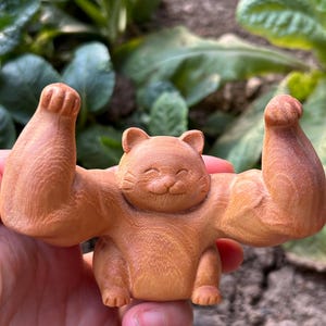 May include: A carved wooden cat figurine with muscular arms raised in a flexing pose. The cat has a smiling face and is made of light brown wood. The figurine is a unique decorative item.