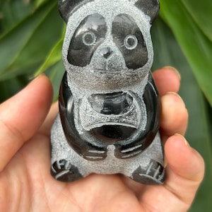 May include: A carved panda figurine with a black and gray color scheme. The panda has black markings around its eyes, ears, and limbs. The figurine is held in a hand, showcasing the detailed craftsmanship of the sculpture.