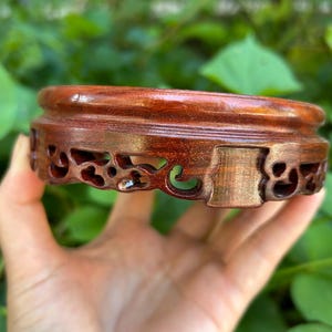May include: A circular, dark brown wooden display stand with detailed carved openwork on its sides. The stand has a smooth, polished finish and is designed to hold a small item. The background features blurred green foliage.