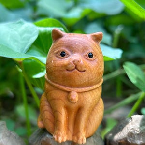 May include: A carved wooden cat figurine with a smiling expression. The cat is light brown with dark eyes and a collar. It is sitting on a wooden post with green foliage in the background. The figurine is approximately 3 inches tall.