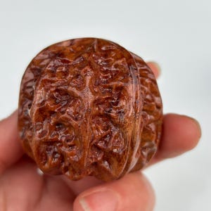 Puede incluir: Una nuez de madera tallada a mano con una superficie detallada y texturizada. La nuez es de un color marrón cálido y rico y parece pulida, lo que resalta las intrincadas tallas. La imagen es un primer plano que muestra la artesanía.