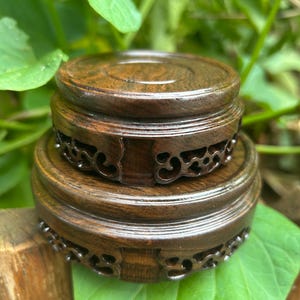 Puede incluir: Dos soportes de exhibición de madera marrón oscuro, uno apilado encima del otro. Los soportes tienen forma circular con patrones decorativos tallados en los lados. Los soportes se colocan sobre un fondo de hojas verdes.