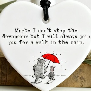 Op de afbeelding: Wit hartvormig keramisch ornament met een zwart koord. Het ornament bevat een citaat: "Misschien kan ik de stortbui niet stoppen, maar ik zal altijd met je meegaan voor een wandeling in de regen." Er is ook een cartoonillustratie van een beer en een varken onder een rode paraplu.